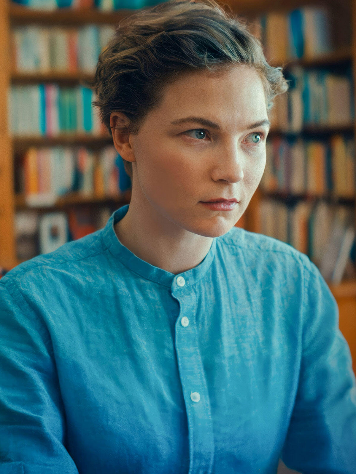 Young woman with short hair in a turquoise shirt, gazing intently to the side while standing in front of a blurred bookshelf background.