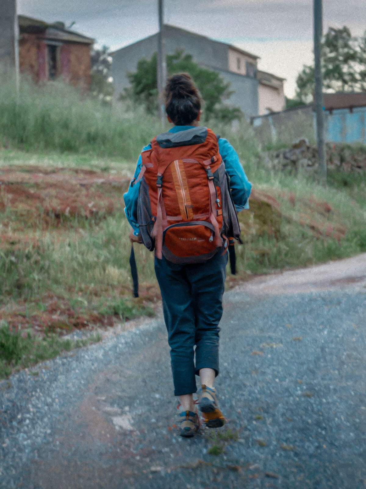 Woman walking alone on a rural road with a large orange hiking backpack, surrounded by overgrown grass and distant village houses.