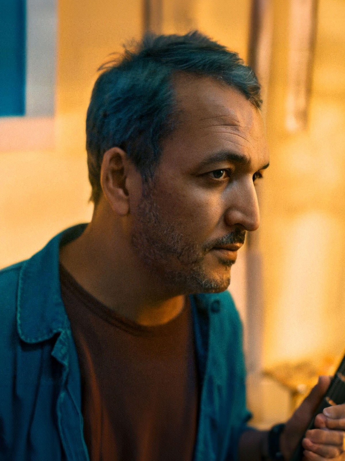 Close-up portrait of a middle-aged man with short hair and stubble, wearing a teal shirt and looking intently to the right, lit by warm indoor lighting.