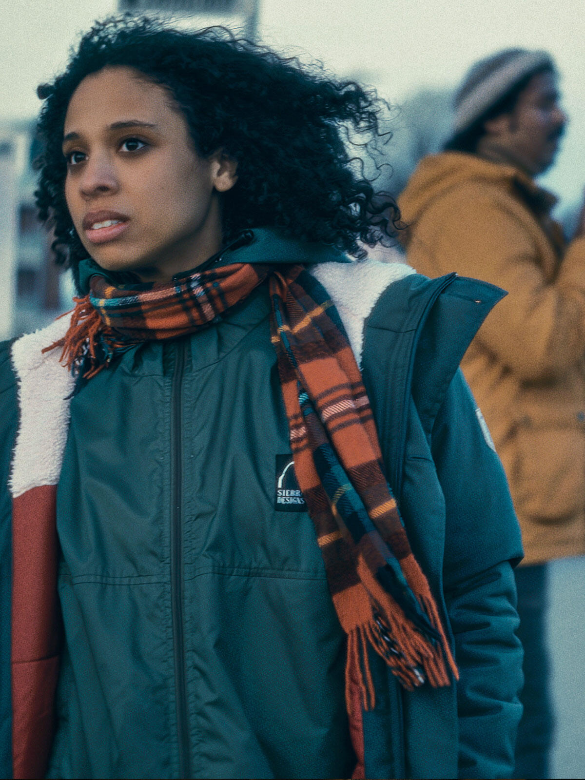 Young woman with curly hair and a red plaid scarf walking outdoors in winter, looking ahead with concern, as a man in a yellow jacket stands blurred in the background.